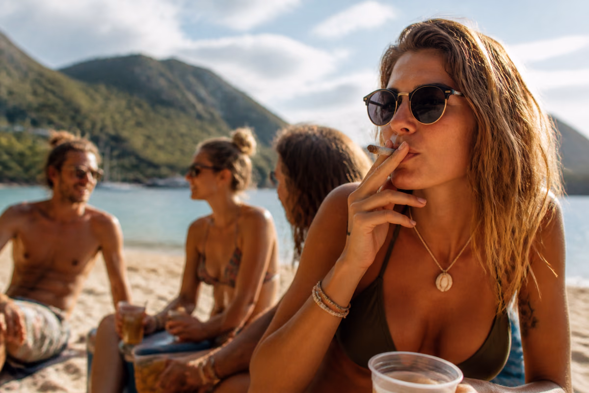 Donna con occhiali da sole fuma uno spinello su una spiaggia tropicale, amici con drink sullo sfondo